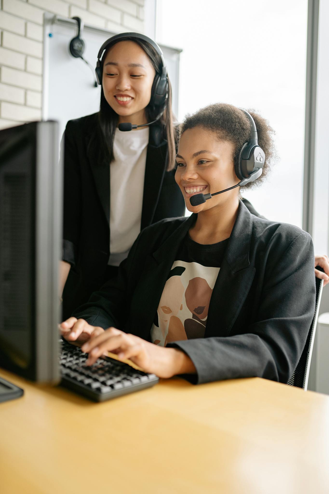 Laughing call center agents wearing headsets collaborate on a computer task in a modern office.